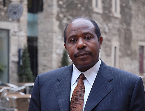 Paul Rusesabagina looking into the camera with a straight face wearing a black suit with a red tie. Photo taken prior to his kidnapping by the Rwandan government.