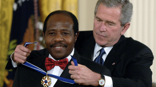 Paul Rusesabagina smiles while wearing a black tuxedo with red bowtie as President George Bush Jr. gives him the Medal of Freedom for his humanitarian work during the Rwandan Genocide.