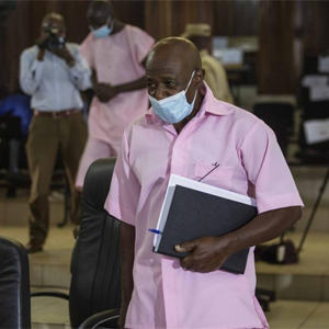 A photo of Paul Rusesabagina in a pink collared shirt with a face mask on, Paul carries a notebook with papers in it as he walks out of the courtroom in Rwanda.