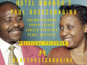 Paul Rusesabagina and his wife smile together, Paul is wearing a pink suit with a brown tie and his wife is wearing a brown dress. Overlayed on the photo is the text "Hotel Rwanda's Paul Rusesabagina, Loving Husband, Proud Father, Cancer Survivor, Peace Activist, Political Prisoner, #FreeRusesabagina"