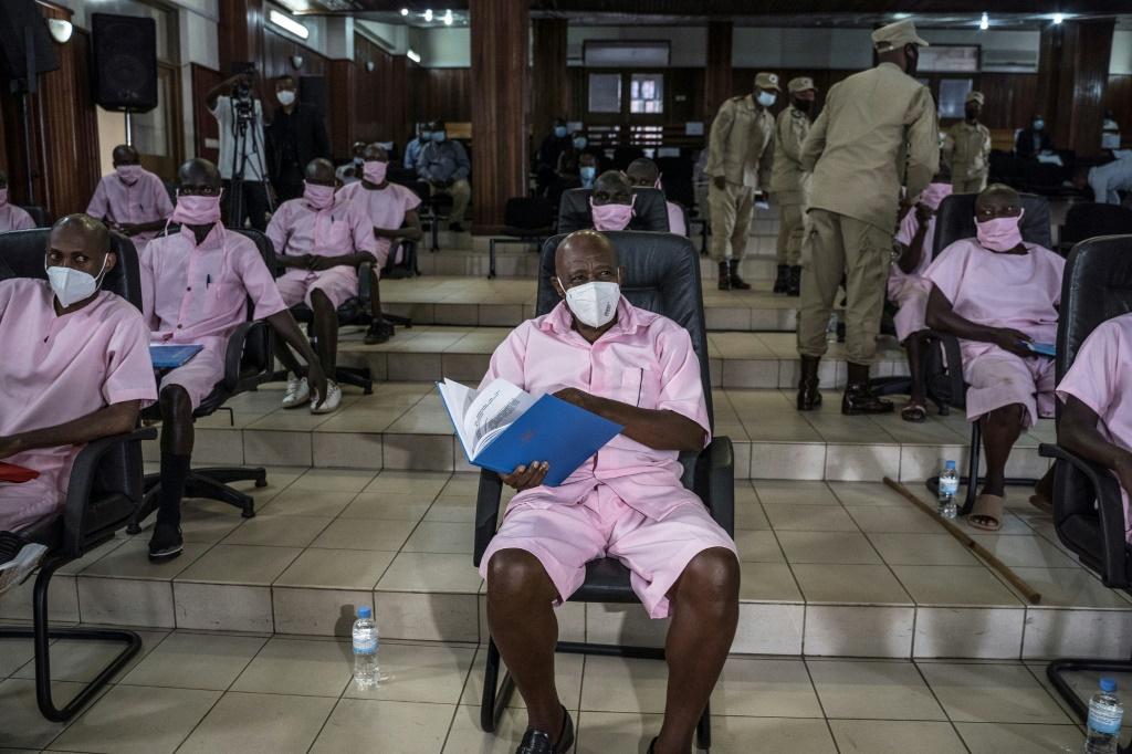 A photo of Paul Rusesabagina in a pink collared shirt and shorts with a face mask on, Paul holds a notebook with papers in it as he sits in the courtroom in Rwanda.