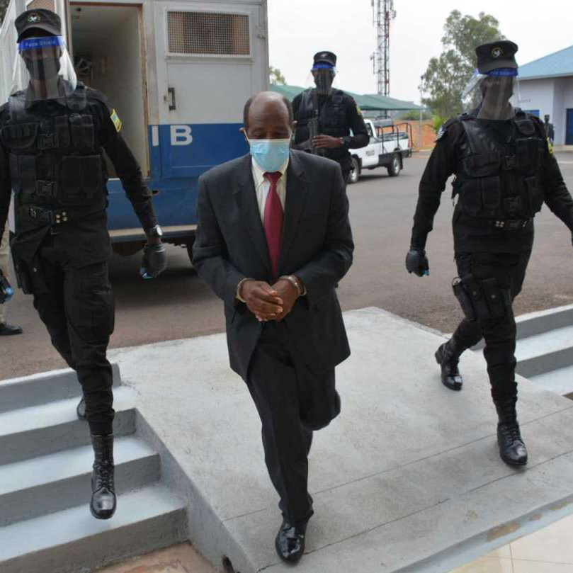 Paul Rusesabaina in a suit and red tie wearing a face mask surrounded by three prison guards in Rwanda in black armed attire walking him into the courtroom.