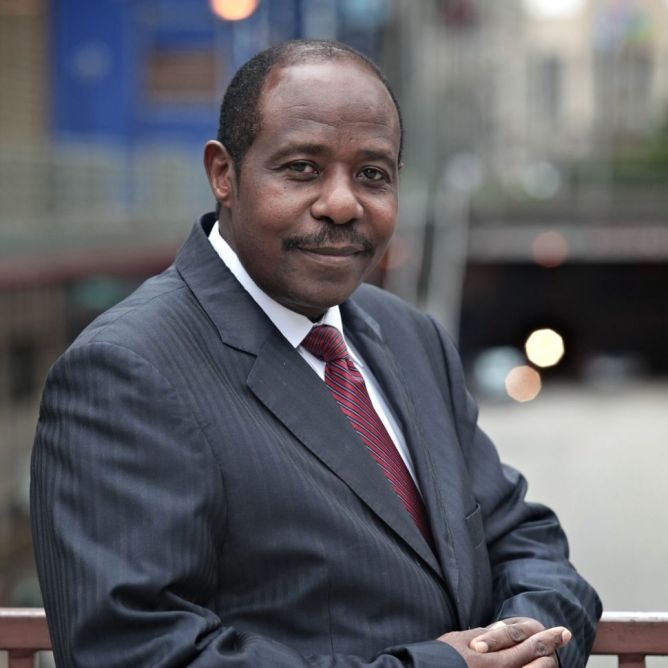 Paul Rusesabagina smiling with his hands clasped wearing a black suit with a red diagonally striped tie. Photo taken prior to his kidnapping by the Rwandan government.